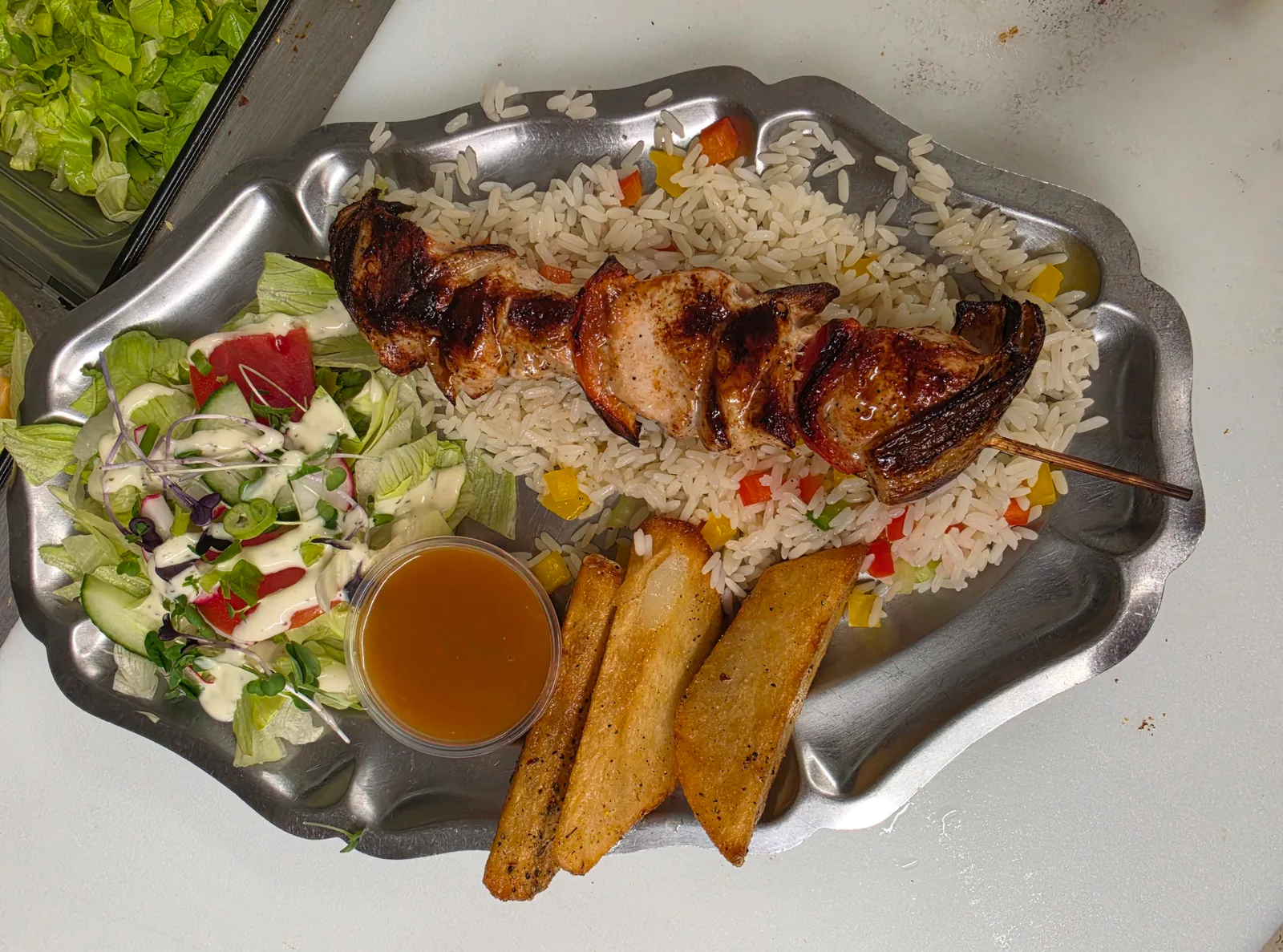 Brochette de viande grillée sur riz aux poivrons, avec quartiers de pommes de terre, salade et sauce, sur un plat en métal
