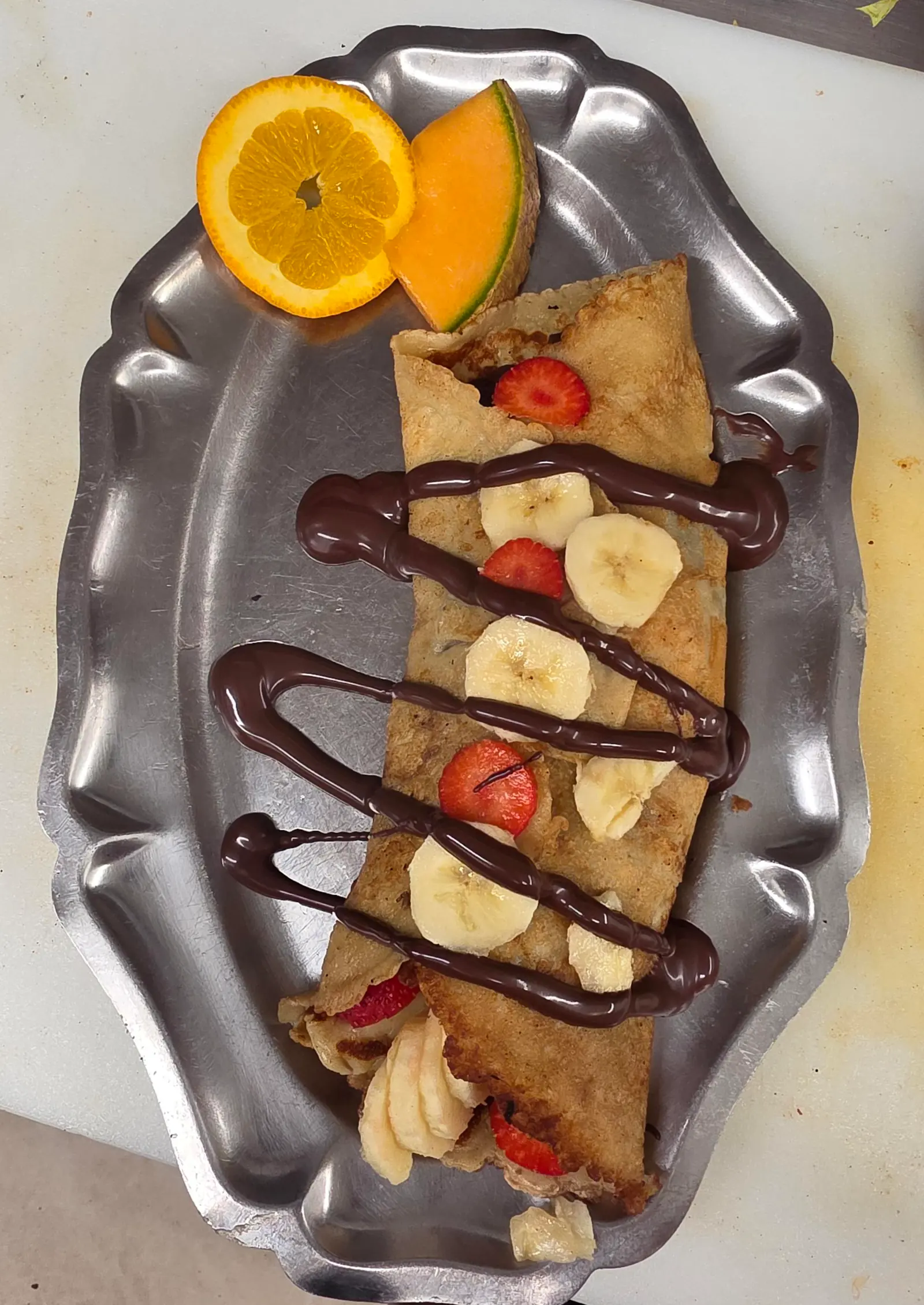 Crêpe garnie de bananes et de fraises, avec un filet de chocolat, servie avec une tranche d'orange et du cantaloup