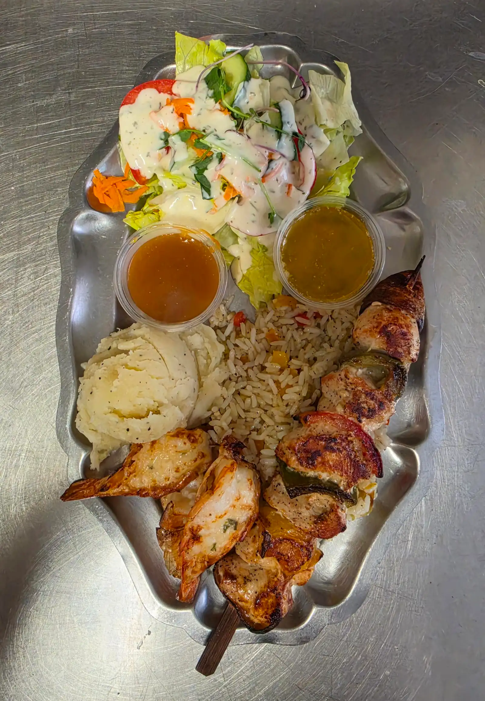 Crevettes grillées, brochette de viande, riz aux légumes, purée et salade avec deux sauces, sur un plat en métal