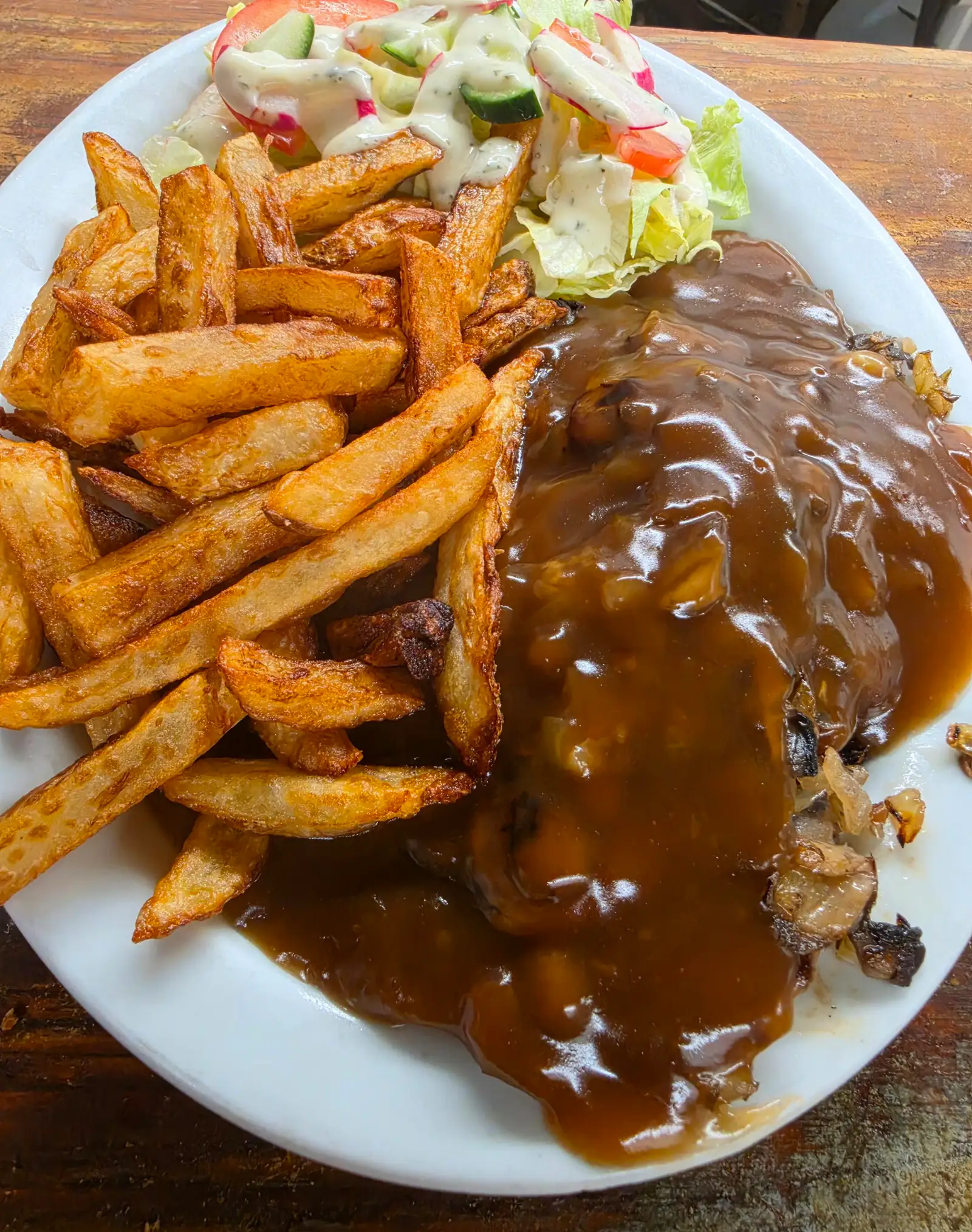 Frites épaisses et portion nappée de sauce brune aux champignons et oignons, avec une petite salade, dans une assiette blanche