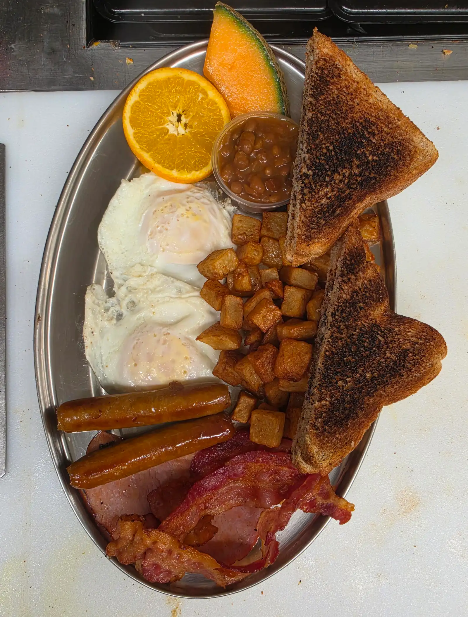 Deux œufs au plat, saucisses, jambon, bacon, patates en cubes, pain grillé, fèves et fruits sur un plat ovale en inox