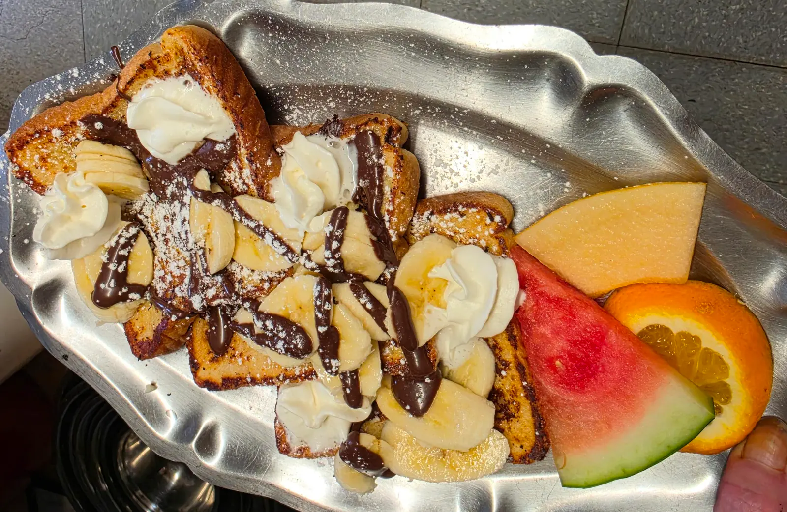 Pain doré garni de bananes, de crème fouettée et d'un filet de chocolat, avec melon d'eau, cantaloup et orange