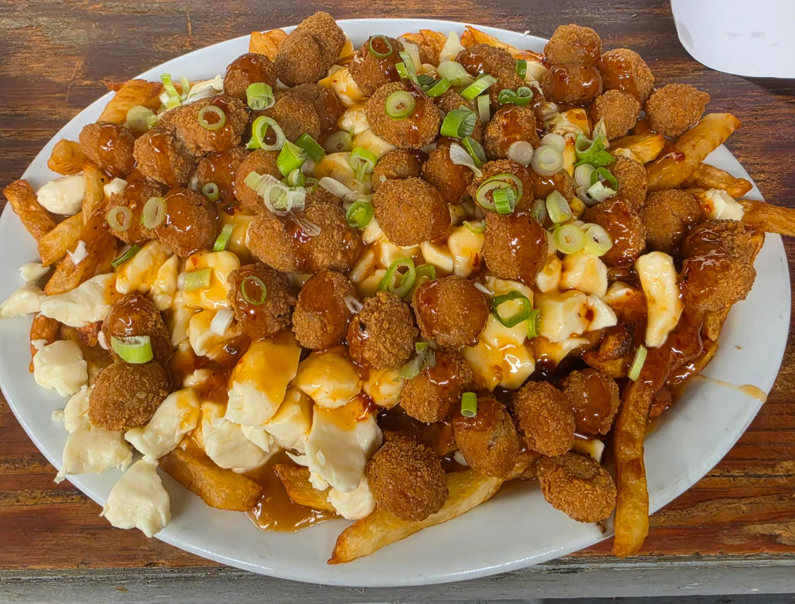 Poutine garnie de bouchées panées, d'une sauce luisante rouge-brun et d'oignon vert tranché, dans une assiette blanche